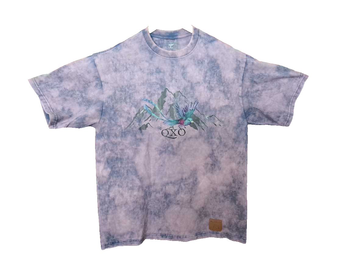 A short-sleeve tie-dye t-shirt in shades of lavender and blue, featuring a central graphic of mountains, tropical leaves, and the OXO logo, with a small brown leather patch near the bottom right hem.