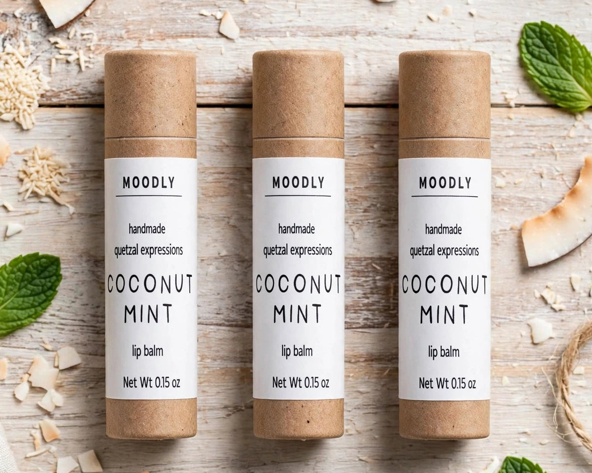 Three MOODLY Coconut Mint lip balm tubes arranged on a rustic whitewashed wooden surface with scattered coconut pieces, mint leaves, and natural fiber accents.